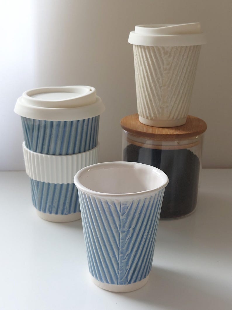 May include: A collection of ceramic coffee cups and travel mugs. The cups feature a textured, ribbed design in white and blue. One mug is stacked on top of two others. A clear glass jar with a wooden lid is also present.