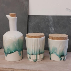 May include: A collection of three ceramic kitchen storage items. There is a tall bottle with a cork stopper and two cylindrical jars, also with cork lids. The containers are white with a teal glaze that drips down the sides.
