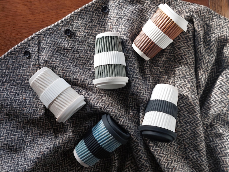 May include: An assortment of reusable coffee cups in various colors, including white, brown, blue, and gray. Each cup features a ribbed design and a contrasting silicone band for grip. The cups are arranged on a textured gray fabric.
