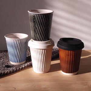 May include: Four ceramic coffee cups with textured, vertical ribbed designs. The cups are in shades of blue, white, brown, and black. One cup has a black lid. The cups are arranged on a wooden surface with a patterned textile.