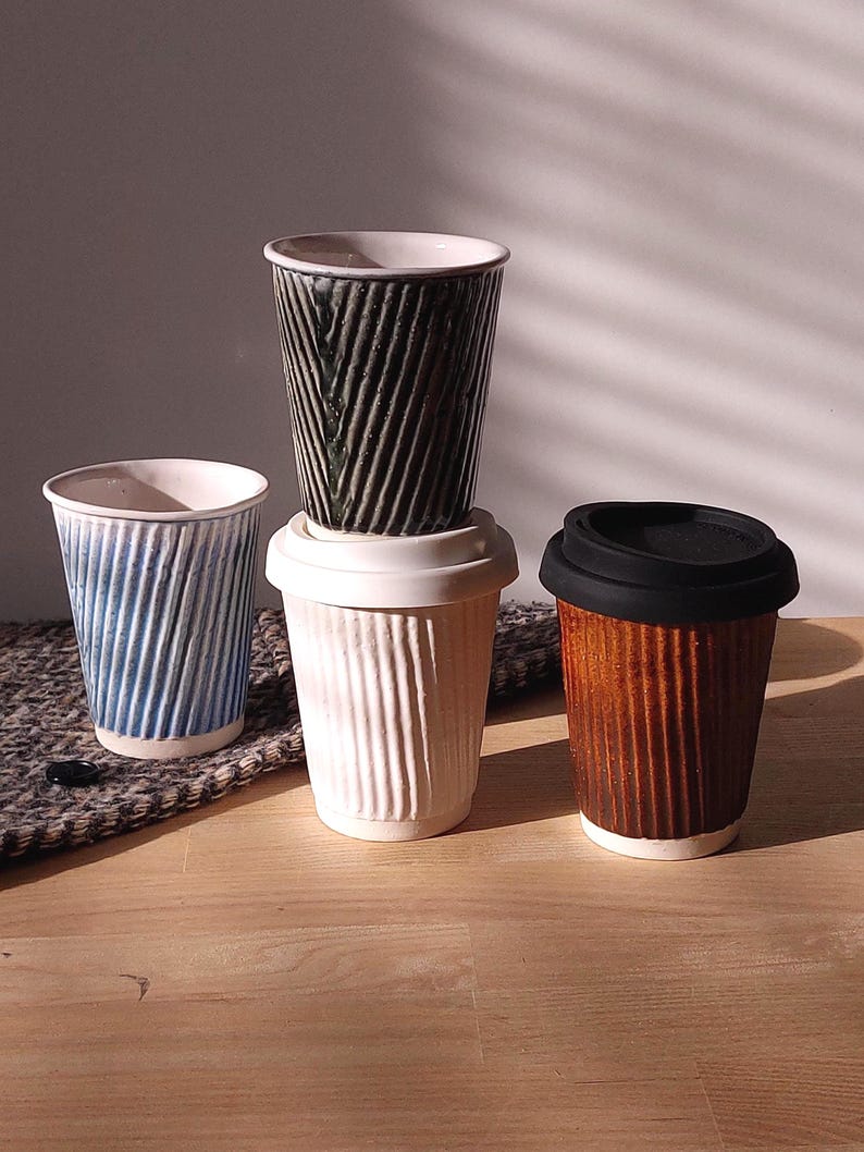 May include: Four ceramic coffee cups with textured, vertical ribbed designs. The cups are in shades of blue, white, brown, and black. One cup has a black lid. The cups are arranged on a wooden surface with a patterned textile.