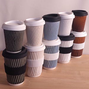 May include: A collection of ceramic travel mugs with textured, ridged exteriors. The mugs are in various colors, including black, white, blue, and brown, with matching or contrasting lids and bands. The mugs are stacked on a wooden surface.