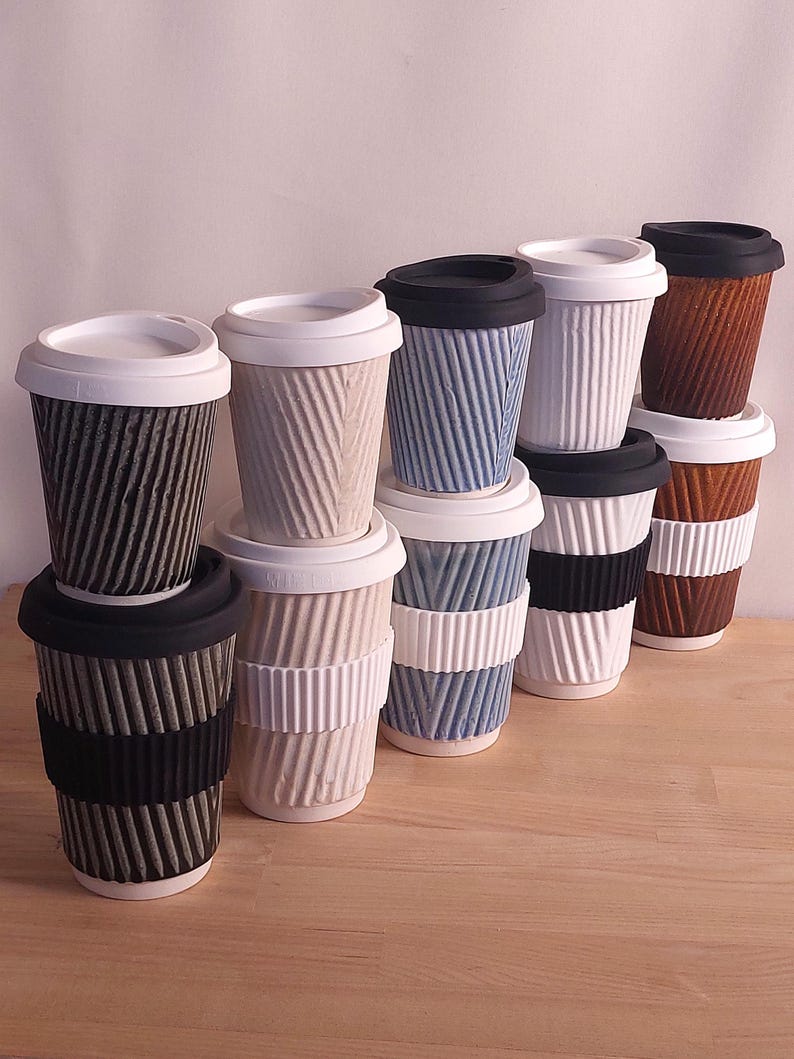 May include: A collection of ceramic travel mugs with textured, ridged exteriors. The mugs are in various colors, including black, white, blue, and brown, with matching or contrasting lids and bands. The mugs are stacked on a wooden surface.