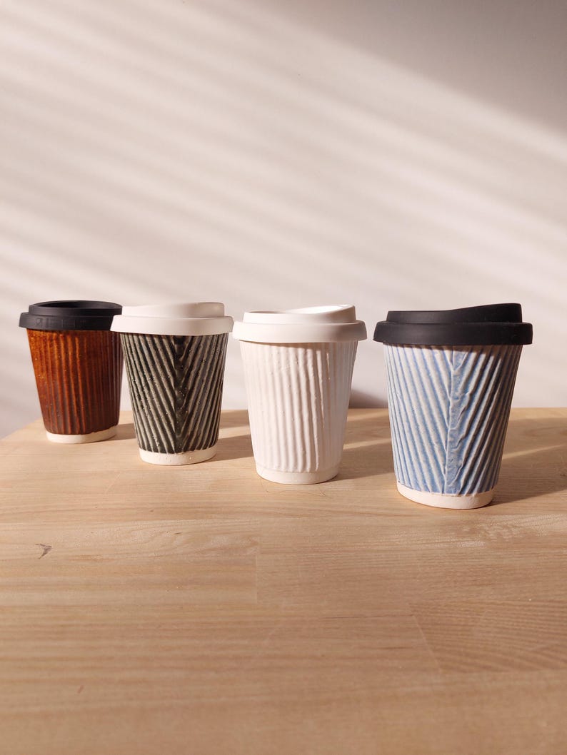 May include: Four ceramic coffee cups with lids. The cups are in various colors: brown, black and white, white, and blue. Each cup has a textured, ribbed design and a contrasting lid.