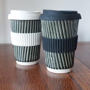 May include: Two ceramic travel mugs with ribbed, olive-green exteriors. One has a white lid and band, the other a black lid and band. Both have white bases and are designed for hot beverages. The mugs are on a wooden surface.