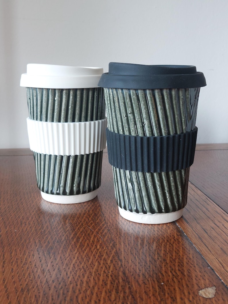 May include: Two ceramic travel mugs with ribbed, olive-green exteriors. One has a white lid and band, the other a black lid and band. Both have white bases and are designed for hot beverages. The mugs are on a wooden surface.