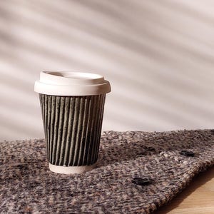 May include: A disposable coffee cup with a white lid. The cup is dark green with vertical ridges and sits on a textured, gray fabric with black buttons. The background is a plain white wall.