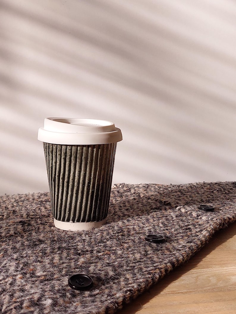 May include: A disposable coffee cup with a white lid. The cup is dark green with vertical ridges and sits on a textured, gray fabric with black buttons. The background is a plain white wall.