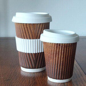 May include: Two brown ceramic coffee cups with white lids. The larger cup has a white ribbed sleeve. The cups have a textured, corrugated design. The cups are on a wooden surface.