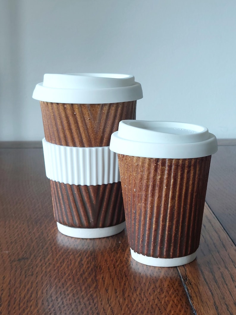 May include: Two brown ceramic coffee cups with white lids. The larger cup has a white ribbed sleeve. The cups have a textured, corrugated design. The cups are on a wooden surface.