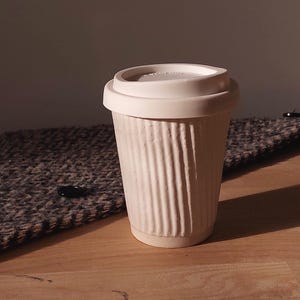 May include: A white ceramic travel mug with a ribbed exterior and a matching lid. The mug is designed to hold hot or cold beverages. The mug is sitting on a wooden surface.