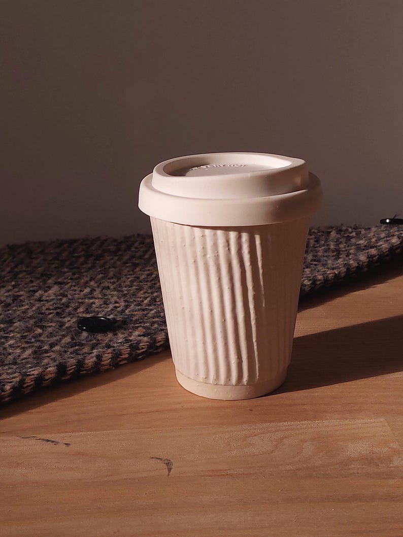 May include: A white ceramic travel mug with a ribbed exterior and a matching lid. The mug is designed to hold hot or cold beverages. The mug is sitting on a wooden surface.