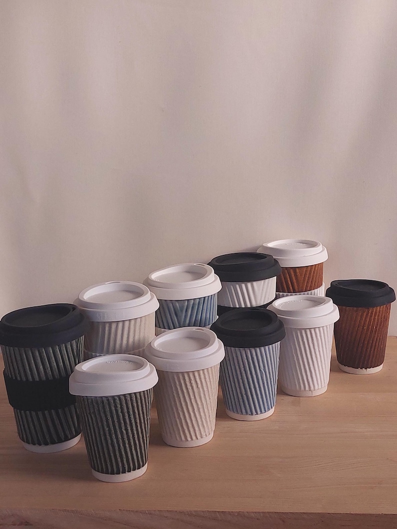 May include: A collection of ceramic coffee cups with lids. The cups have a ribbed texture and come in various colors, including white, black, blue, and brown. Some cups have black silicone sleeves. The lids are white or black.