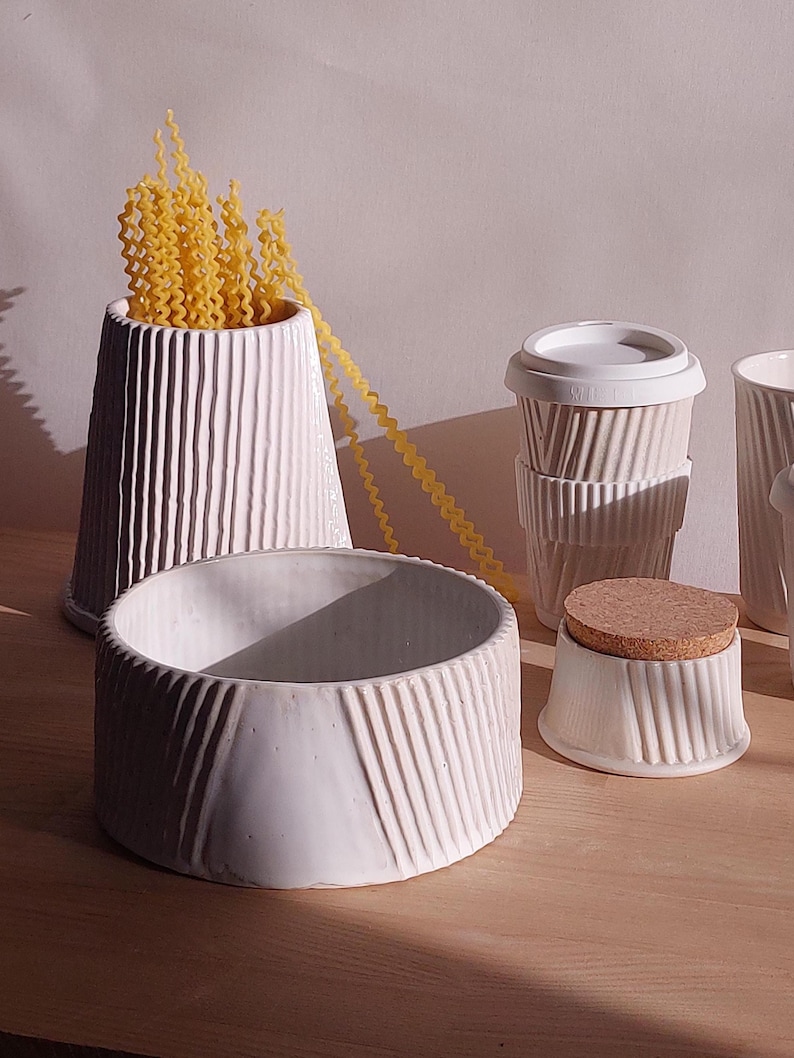May include: A collection of white ceramic kitchenware with a ribbed design. Includes a vase filled with yellow pasta, a bowl, a small jar with a cork lid, and two stacked cups with lids. The items are arranged on a wooden surface.