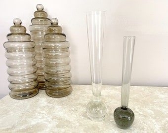 May include: A collection of decorative glass vases and jars. The jars are a smoky gray color with a stacked, rounded design. Two tall, clear vases have a bulbous base, and a smaller vase has a dark gray base.