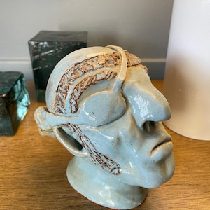 May include: A blue ceramic sculpture of a person's head with brown details. The sculpture has a large nose and a closed mouth. The person is wearing a headband with a brown design.