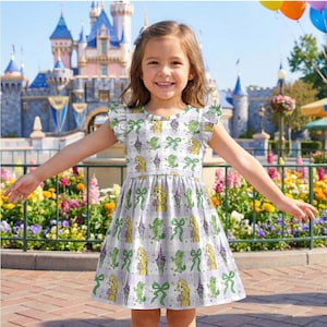 May include: A young girl wearing a white dress with a Rapunzel-themed print, featuring images of the character, bows, and towers. The dress has short, ruffled sleeves and a flared skirt. The background includes a castle, flowers, and balloons.