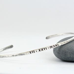 May include: A silver hammered bangle bracelet inscribed with the date "VIII. XXVI. MMXI".
