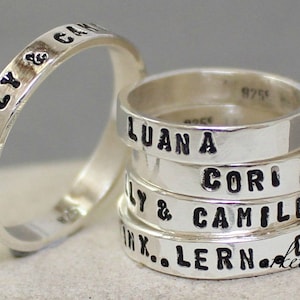 Könnte beinhalten: Ein Stapel aus fünf silbernen Ringen mit schwarzer Gravur. Die Ringe sind mit den Namen "Lilly & Cam", "Luan A", "Cori", "Ly & Camille" und "Ink... Lern 00..." graviert.