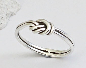 Op de afbeelding: Een zilveren knoop ring met een eenvoudig, elegant ontwerp. De ring is gemaakt van één stuk metaal dat is gedraaid en geknoopt om een unieke en opvallende look te creëren.