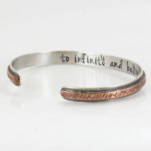 Könnte beinhalten: Ein silbernes Armband mit einem kupfernen Band, auf dem die Worte "bis in die Unendlichkeit und darüber hinaus" eingraviert sind.