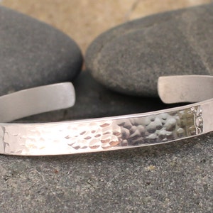 May include: A silver cuff bracelet with a hammered texture on the front and a smooth, polished finish on the inside. The open-ended design allows for easy wear. The bracelet is displayed on a gray stone surface.