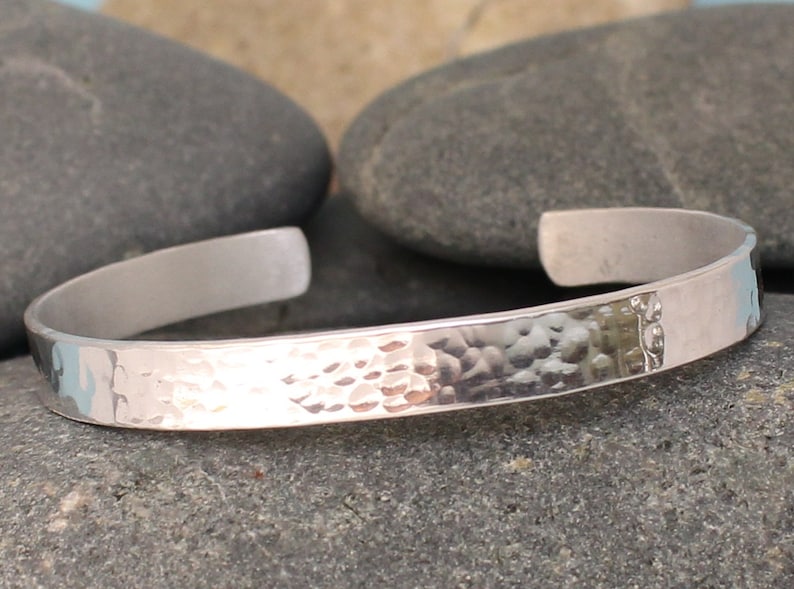 May include: A silver cuff bracelet with a hammered texture on the front and a smooth, polished finish on the inside. The open-ended design allows for easy wear. The bracelet is displayed on a gray stone surface.