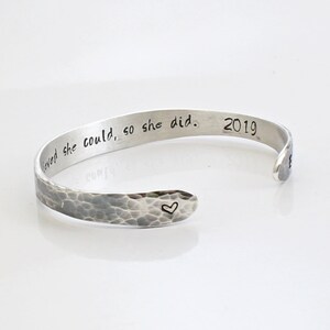May include: Silver-toned cuff bracelet with a hammered texture. The bracelet features the inscription "believed she could, so she did. 2019" and a small heart symbol. A simple, elegant piece of jewelry.