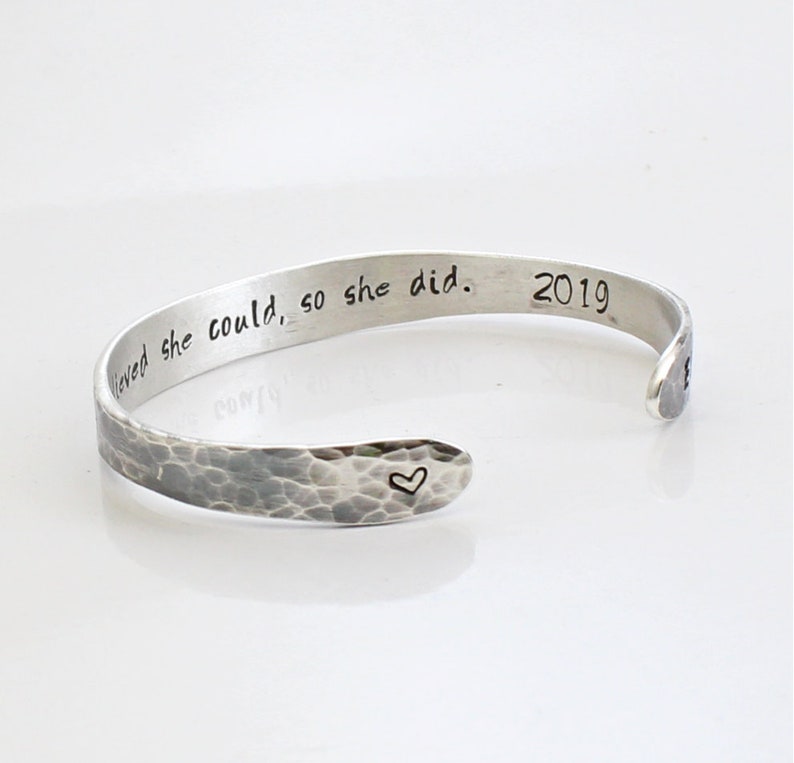 May include: Silver-toned cuff bracelet with a hammered texture. The bracelet features the inscription "believed she could, so she did. 2019" and a small heart symbol. A simple, elegant piece of jewelry.