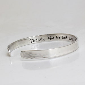 May include: A silver cuff bracelet with the inscription "Though she be but little she is fierce." The bracelet has a hammered texture on the lower half and a smooth, polished finish on the upper half.