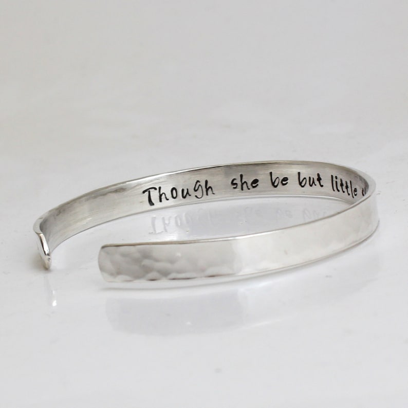 May include: A silver cuff bracelet with the inscription "Though she be but little she is fierce." The bracelet has a hammered texture on the lower half and a smooth, polished finish on the upper half.