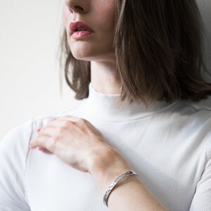 May include: A silver hammered cuff bracelet on a wrist. The bracelet has a textured surface with a repeating pattern. The person is wearing a white turtleneck sweater.