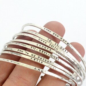 May include: A stack of silver bangles with various personalised inscriptions, including "Love you more", "12.11.2017", "Have faith", "Sophie", "VII. XXVI. MMXI", "You're my person", "Katie", and "AHE".