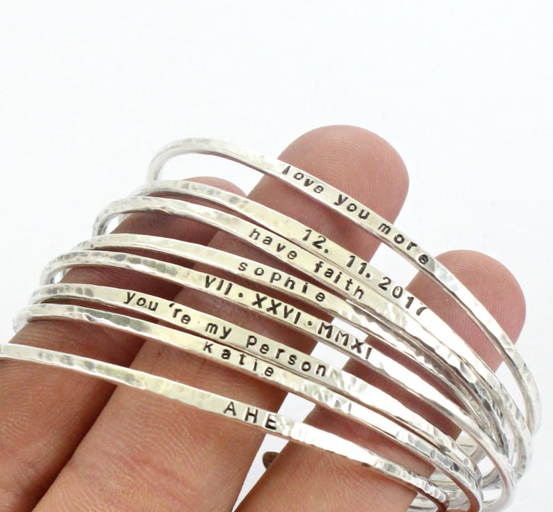 May include: A stack of silver bangles with various personalised inscriptions, including "Love you more", "12.11.2017", "Have faith", "Sophie", "VII. XXVI. MMXI", "You're my person", "Katie", and "AHE".