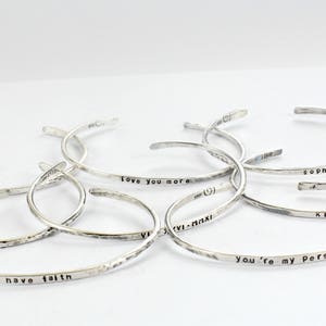 May include: A collection of silver bracelets with different engraved messages, such as "Love you more", "Sophie", "Katie", "You're my person", "12. 11. 2", "AHE", and "have faith".