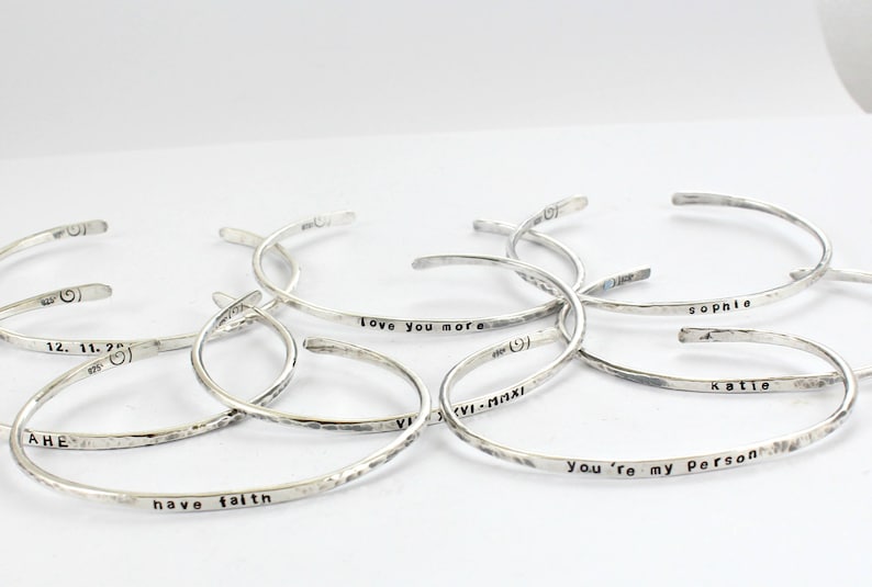 May include: A collection of silver bracelets with different engraved messages, such as "Love you more", "Sophie", "Katie", "You're my person", "12. 11. 2", "AHE", and "have faith".