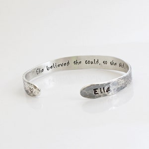 May include: A silver-toned cuff bracelet with a hammered texture. The bracelet is engraved with the words "She believed she could, so she did." and the name "Ella." A small heart is stamped near the opening.