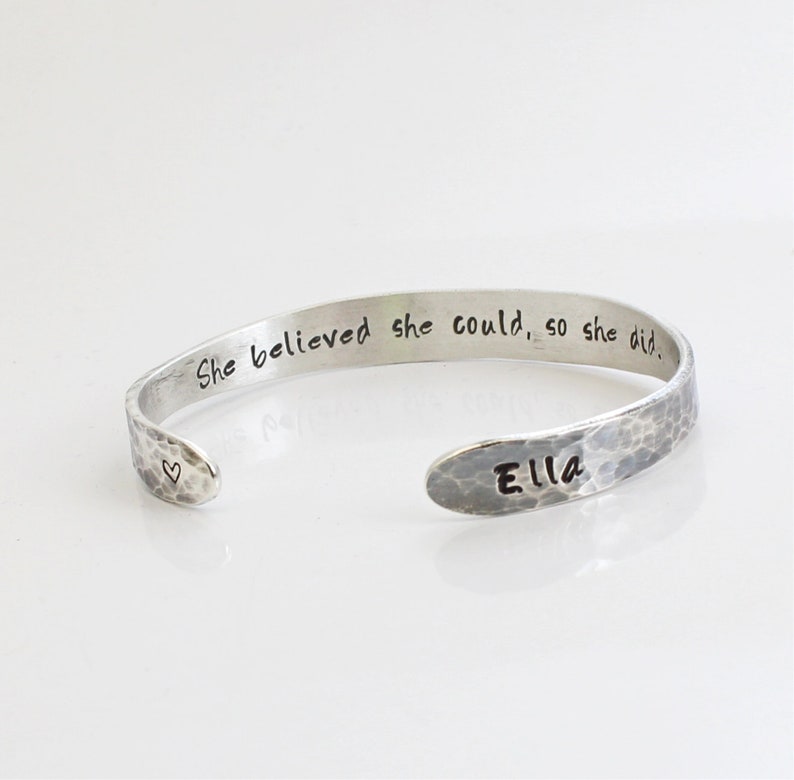 May include: A silver-toned cuff bracelet with a hammered texture. The bracelet is engraved with the words "She believed she could, so she did." and the name "Ella." A small heart is stamped near the opening.