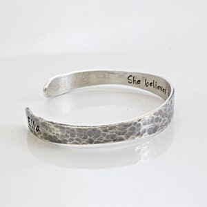May include: A silver cuff bracelet with a hammered texture. The bracelet is engraved with the words "Ella" and "She believed". The open-ended design allows for easy wear. The bracelet is displayed on a white surface.