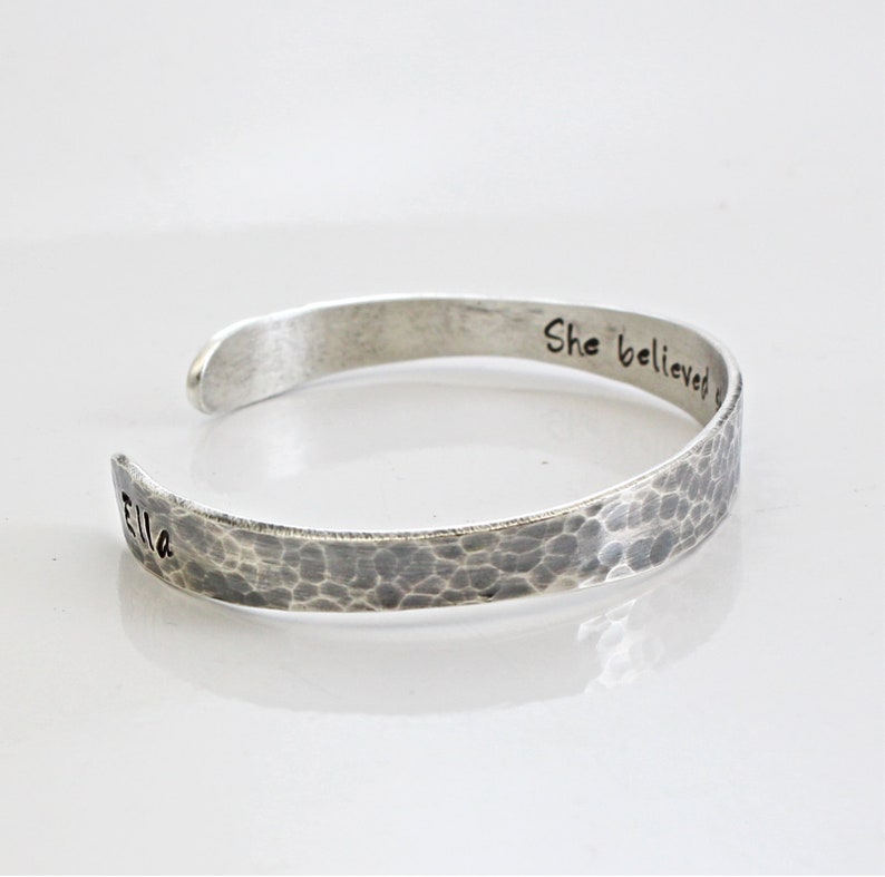May include: A silver cuff bracelet with a hammered texture. The bracelet is engraved with the words "Ella" and "She believed". The open-ended design allows for easy wear. The bracelet is displayed on a white surface.