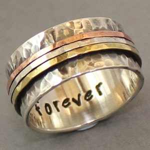 Op de afbeelding: Een draaibare zilveren ring met drie banden van koper, messing en zilver. De ring is gegraveerd met het woord "forever".