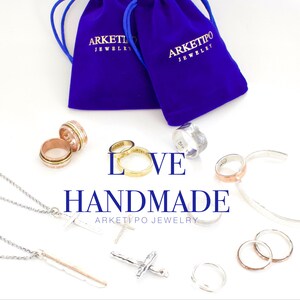 May include: A collection of handmade jewellery, including rings, necklaces, and bracelets, displayed on a white surface. The jewellery is made of silver, gold, and copper. The image also includes two blue velvet bags with the text "Arketipo Jewellery" printed on them. The text "LOVE HANDMADE ARKETIPO JEWELLERY" is also displayed on the surface.