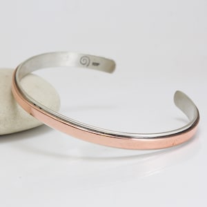 May include: A silver cuff bracelet with a copper-colored band. The bracelet has a simple, open design and is displayed on a white surface. The metal cuff is approximately 2.5 inches in diameter.