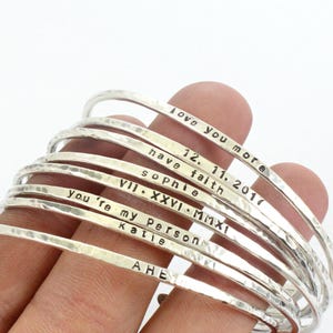 May include: A stack of silver bangle bracelets with various engraved messages, including "love you more", "have faith", "sophie", "VII. XXVI. MMXI", "you're my person", "Katie", and "AHE".