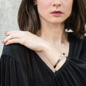 May include: A hammered metal bracelet with a black band is worn on a wrist. The bracelet has a textured, reflective surface. The person is wearing a black pleated top and has red nail polish. Small gold hoop earrings are visible.