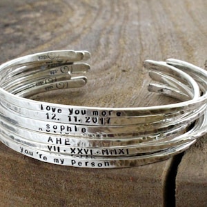 Könnte beinhalten: Ein Stapel silberner Armbänder mit personalisierten Gravuren. Die Gravuren beinhalten "love you more", "12. 11. 2017", "Sophie", "AHE", "VII. XXVI. MMXI" und "You're my Person".