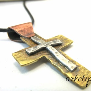 Puede incluir: Un colgante de cruz de plata y latón con textura martillada, colgado de un cordón negro. La cruz está unida a una baíl de color cobre.