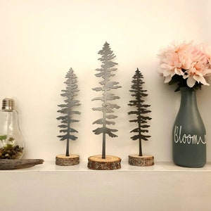 May include: Three metal tree silhouettes on wooden bases, a grey vase with the word "Bloom" written on it, and a glass lightbulb with a small plant inside. The trees are different sizes and are arranged on a white shelf.