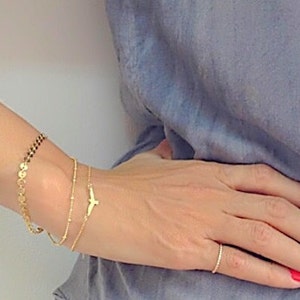 Può includere: Un polso di donna con tre braccialetti d'oro. Il braccialetto superiore è una catena con piccoli anelli rotondi. Il braccialetto centrale è una catena delicata con un piccolo ciondolo a forma di uccello. Il braccialetto inferiore è una catena sottile.