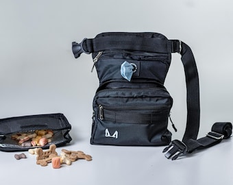 May include: Black dog walking bag with multiple compartments, including a dispenser for waste bags, a pouch for treats, and a zippered pocket. The bag has an adjustable strap and a logo that says "M".